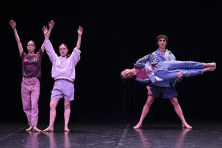 Vier Tänzer*innen auf der Bühne: Zwei links im Foto mit den Armen weit nach oben gestreckt. Die anderen beiden rechts. Hier trägt einer den anderen. Dieser streckt sich horizontal