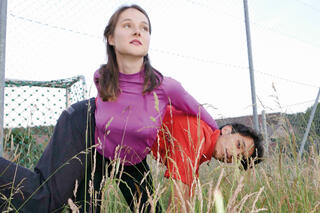 Jugendliche in hohem Gras vor einem Stacheldrahtzaun, sie hält ihn quer auf dem Rücken
