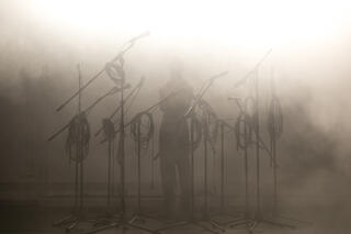 Der Performer Tiran Willemse ist von dichtem Nebel und Mikrofonständern umgeben, deren Kabel aufgerollt sind. Seine beiden Arme hält er in einer Umarmung dicht an seinem Körper.