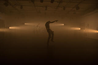 Der Performer Tiran Willemense ist von Nebel umgeben und wird von gelbem Licht angestrahlt. Seinen Körper siet man nur schemenhaft.