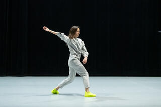 Eine weiblich gelesene Tänzerin steht auf einer weißen Bühne mit schwarzem Hintergrund. Sie ist weiß gekleidet, steht in einem Ausfallschritt und hat ihren vorderen Arm nach hinten gestreckt.