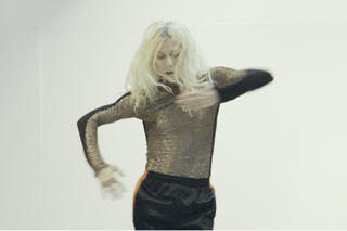 Eine weiblich gelesene Person tanzt und bewegt dabei ihre Arme, das Bild ist leicht verschwommen durch die Bewegung.
