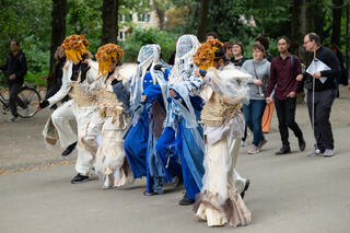 Eine Fotografie der Performance "Erste Schritte zu den Schwellen" von Zwoisy Mears-Clarke, die im Hofgarten stattfand. Im Vordergrund laufen fünf Performer*innen nebeneinander über einen Weg, der von Bäumen umgeben ist. Sie tragen Kostümen aus fließenden Stoffen und Masken, die ebenfalls aus Stoffen bestehen. Ihre Arme sind ineinander eingehakt. Im Hintergrund ist eine Gruppe von Personen, die der Performance beiwohnt und den Weg der Performer*innen verfolgt. Zwei der Personen tragen einen Langstock.