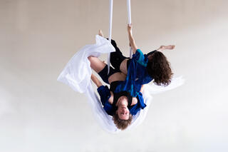 Zwei Personen hängen gemeinsam zwei Vertikaltüchern (Silks). Eine der Personen hängt über Kopf und die andere zur Seite.