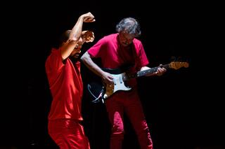 Auf dem Foto ist ein Performer hinter jemandem zu sehen, der Gitarre spielt.