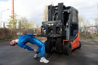 Ein/e Performer*in in einem blauen Einteiler steht vor einem Gabelstapler. Die Person hält sich an der Ladefläche fest und beugt sich dabei nach hinten. 