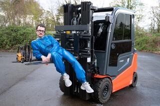 Ein/e Performer*in sitzt, schräg angelehnt, auf der Ladefläche eines Gabelstaplers. Die Person trägt einen blauen Einteiler.