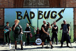 Die Band "Bad Bugs" steht vor einem Banner mit ihren Instrumenten, auf dem ihr Name geschrieben ist. Sie tragen alle schwarze Kleidung.