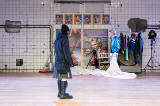 Zwei Personen stehen in einer gefliesten Halle. Sie schauen eine Wand an, an der verschiedene Portraitfotos von Personen hängen. Ebenso hängen große Ausdrucke von diversen Magazincovern an der Wand. Daneben steht eine Kleiderstange mit verschiedenen Kleidungsstücken.