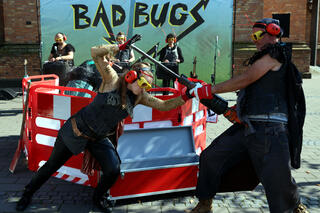 Die Band Bad Bugs in einer "Baustellen-Szene": vorne agieren zwei miteinander, im Hintergrund vor einer Backsteinwand und einem Bad Bugs Banner drei Bandmitglieder*innen an Instrumenten