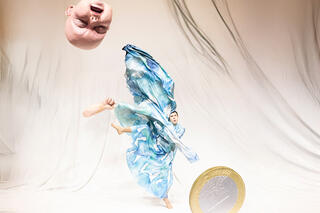 Eine KI-Installations-Collage eines Babykopfes, einer 1-Euro-Münze, eines Unterarmes mit Hand und eines Mannes in Tanzpose mit engelhaft schwingender, blau-weißer Stoffbekleidung