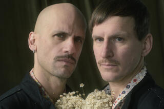 Ein Portraitfoto des Choreografen-Duos HARTMANNMUELLER . Sie stehen dicht beinander und tragen schwarze Hemden. Zwischen ihnen befinden sich Blumen.