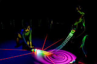 Zwei Performer*innen befinden sich auf einer dunklen Bühne. Das einzige Licht geht von einer regenbogenfarbenen Spielzeugspirale aus, die beide Performer*innen gemeinsam halten. Durch das auf die Spirale fallende Licht entsteht ein spiralförmiger Schatten auf dem Bühnenboden. Ebenso ist Klebeband auf dem Bühnenboden, das im Dunkeln leuchtet.