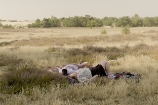 Zwei Menschen liegen auf einer Decke in einem gelben Heide-Feld. Es scheint Sommer zu sein. Sie liegen aneinander und auf dem Rücken. Sie blicken in den Himmel.