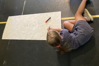 Ein Kind liegt seitlich auf dem Boden, es hält einen Stift in der Hand. Daneben liegt ein weißes Plakat, auf dem das Wort Macht in Großbuchstaben steht.