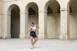 Eine Tänzerin in einer schwarzen, kurzen Hose und einem weiß-schwarzen Top tanzt auf einem Innenhof, der von einem Gebäude umgeben ist. Vom Gebäude ist ein Gang zu erkennen, der zum Innenhof durch Säulen geöffnet ist. Die Tänzerin nimmt eine pliéartige Pose ein, die Beine sind leicht angewinkelt und das rechte nach vorne gerichtet. Die Arme hält sie angewinkelt vor den Oberkörper, während ihr Blick zur Seite gerichtet ist.