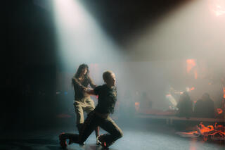 Zwei Personen befinden sich auf einer schwach beleuchteten Bühne. Sie werden von zwei Scheinwerferkegeln angestrahlt. Eine der Personen ist mit dem Rücken zur anderen gewandt. Die hintere Person hält die vordere Person an den Händen, die vordere lehnt sich im Ausfallschritt nach vorne. Beide tragen Trainingshosen und Oberteile aus Spitzenmaterial.