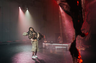 Drei Personen befinden sich auf einer rot beleuchteten Bühne. Im Hintergrund sitzt eine Person vor einem flachen Mischpult und singt in ein Mikrofon. Die beiden anderen Personen befinden sich im vorderen Bereich der Bühne. Die eine Person trägt die andere Person in den Armen. Ihre Augen sind leicht geschlossen. Beide tragen Trainingshosen sowie ein Oberteil aus Spitzenmaterial. Im rechten Bildrand sieht man eine Textilinstallation aus rotem Stoff.