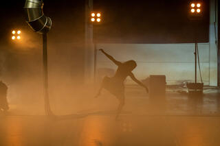 Eine Bühne ist mit Nebel gefüllt, der von schwachen Lichtern gelb-orange angestrahlt wird. Inmitten des Nebels befindet sich die Silhouette eines*R Tänzer*in der*die die Arme gerade zur Seite streckt und das rechte Bein angewinkelt hat. Der Körper befindet sich in einer leicht zur Seite geneigten Position. Von links oben ragt ein silbernes Rohr, von dem aus lange, schwarze Seile auf den Boden reichen, in das Bild.