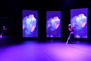 Laura Waltz steht auf einer lila-beleuchteten Bühne. Vor ihr drei Screens die ihre Bewegung in farbenfrohe Striche malt.