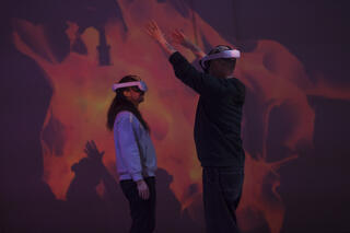 Zwei Personen stehen sich gegenüber und tragen VR-Brillen, während eine von ihnen die Arme hebt. Der Hintergrund hat eine düstere Atmosphäre.