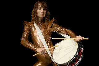 Eine Person in einem glänzenden, bronzefarbenen Ganzkörperanzug trägt eine Trommel mit einem Gurt, auf der sie mit zwei Drumsticks spielt. Der Anzug liegt eng an, darunter ist eine Attrappe angebracht, sodass die Person schwanger wirkt.