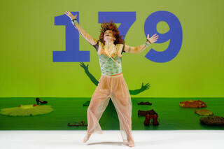 Performerin mit rot-braunen Locken auf der Bühne und streckt die Arme aus. Sie trägt eine weiße durchsichtige Flatterhose, darunter eine beige Leggins. Als Oberteil ein durchsichtiges Hemd, darunter ein türkises Top mit gelb-floresken Elementen. Im Hintergrund ist die Bühne grün erleuchtet und es steht mit großen blauen Ziffern 1 7 9 auf der Wand. Auf dem Boden liegen Kleidungsstücke.