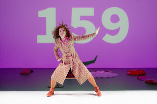 Performerin Liz Santoro steht mit braun-roten Locken auf der Bühne. Sie trägt einen orange-karierten Overall. Hinter ihr eine Lila erleuchtete Bühne auf der in großen Ziffern die Zahlen 1 5 9 stehen. Auf dem Boden hinter ihr liegen Kleidungsstücke wie zum Beispiel einen Tütü.