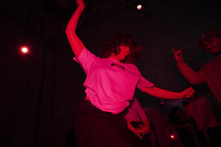 Eine Person mit weißen T-Shirt, schwarzer Hose und mittellangen braunen Haaren ausgelassen tanzend auf der Tanzfläche, erleuchtet in rotem Licht. Um sie herum Menschen, die sie anfeuern.