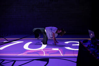 Eine Licht-Installation auf dem Boden. Eine Teilnehmer*in auf dem Boden in tänzerischer Haltung mit einem Bein zur Seite gestreckt. Stützt sich mit den Händen ab. Auf dem Boden die Ziffern 70,3. Im Vordergrund ist ein Pult mit einem Laptop zu sehen.