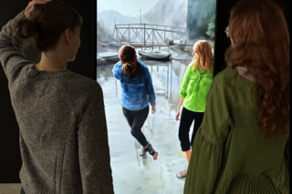 Zwei Menschen mit langen, roten, leicht lockigen Haaren und mit dunkel bruanen Haaren vor einem Screen der sie beide als Avatar malerisch abbildet. Die linke Person hebt die Hand und legt sie auf den Kopf. Im Abbild macht das der Avatar auch. Im Hitnergrund ist eine malerische Landschaft mit kleinen Booten, einer Brücke zu sehen. Ein Gebirge zeichnet sich dahinter ab.