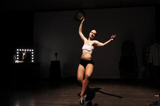 Das Bild zeigt eine Person die iher linkes Bein vor dem anderen hat. Sie streckt beide Arme in die Höhe und hält in ihrer rechten Hand einen Hut. Die Person hat ihre Augen geschlossen. Im Hintergrund ist ein Schminktisch mit beleuchtendem Spiegel zusehen. Die Person wird von einem Scheinwerfer erleuchtet. Das restliche Bild ist dunkel.