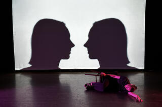 Das Bild zeigt eine Person die rechts im Bild auf dem Boden liegt. Sie liegt auf ihrer rechten Seite, mit dem Kopf in Richtung Publikum. Die Person hat ihren linken Arm ausgestreckt. Die Person trägt dunkle Klamotten. Sie hat außerdem einen Hocker unter sich. Im Hintergrund ist eine Leinwand zusehen auf der Schattenumrisse von zwei Köpfen zusehen sind. Diese gucken sich an.