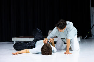 Zwei Personen befinden sich gemeinsam auf einem weißen Bühnenboden. Eine der Personen liegt auf dem Rücken, während die andere neben ihr kniet und ihre Hand auf den Brustkorb der liegenden Person legt. Die liegende Person hält ihren Arm in Richtung der Hand auf ihrem Oberkörper.