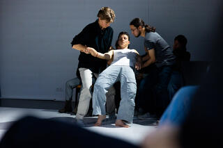 Es ist eine Gruppe von Personen auf einer Bühne zu sehen. Am Rand sitzen Zuschauer*innen, direkt vor ihnen befinden sich drei Performer*innen. Eine Person wird von den anderen beiden seitlich festgehalten, während sie sich mit ihrem Rücken nach hinten fallen lässt.