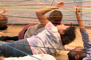 Das Bild zeigt ein Netzt das aus Bunten Fäden gespannt ist. Darunter liegen fünf Kinder die mit jeweils einer Hand an dieses Netz tasten.. 