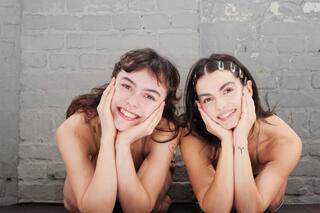 Auf dem Bild sind zwei Personen zu sehen. Beide haben ihren Kopf auf ihren Händen abgestützt und lächeln in die Kamera.