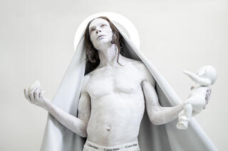 Das Bild zeigt eine Person die oberkörperfrei zusehen ist. Sie ist am gesamten Oberkörper mit weißer Farbe angemalt. Die Person trägt außerdem eine weiße Kopfbedeckung die wie ein Umhang nach unten geht. Sie hält ihre beiden Arme, geknickt nach oben. In der linken Hand hält sie eine komplett weiße Puppe und in der anderen einen Stein. Die Person hat ihren Kopf nach oben gerichtet.