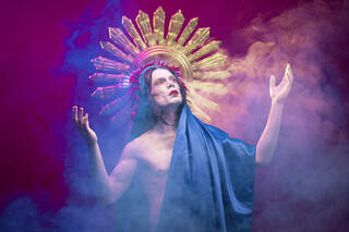 Das Bild zeigt eine Person die etwas zur Seite gedreht steht und dabei nach oben schaut. Sie hat ihre beiden Arme geknickt nach oben gestreckt. Die Person trägt ein blaues Gewand. Im Hintergrund ist ein goldenes Muster zu erkennen. Die Person steht in Nebel.