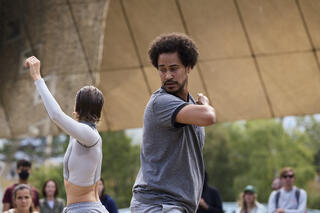 Zwei Tänzer*innen stehen vor einem Publikum in Straßenkleidung in tänzerischer Pose. Sie heben einen Arm in die Höhe. Sie befinden sich unter einem beigen Segeldach, dahinter grüne Flora und die Gesichter von Zuschauer*innen. Es ist Sommer.