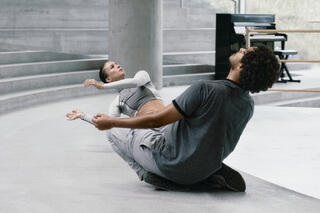 Zwei Tänzer*innen in einer grauen Beton Architektur. Sie sitzen auf den Boden und lehnen die Oberkörper nach hinten. Sie sind in Straßenkleidung. Im Hintergrund steht ein Klavier.