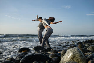 Zwei Performer*innen stehen an der Brandung mit den Füßen im Wasser. Vor ihnen das Meer, hinter ihnen aufgetürmte Steine. Die Tänzer*innen sind in Bewegung und strecken die Arme und Oberkörper nach vorne und hinten. Sie sind miteinander verwebt.