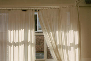 Weißer Vorhang vor Fenster, das zur Seite geschoben wird. Dahinter ist eine Ziegelwand zu sehen. Das Licht leuchtet durch das Fenster.