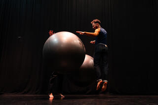 Zwei große silberne Bälle auf einer schwarzen Bühne. Der Perfomer Alfredo Zinola in Jeans und zweifarbigen Shirt springt in die Luft und lässt dabei die Bälle springen. Er lächelt und hat sichtlich Spaß.