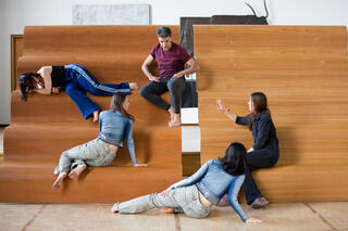 Vier Personen sitzen und liegen verteilt auf einer treppenartigen Skulptur aus Holz. Eine weitere Person liegt davor seitlich auf dem Boden. Eine der Personen rechts spricht, während sie mit der rechten Hand gestikuliert, die anderen richten sich zu ihr.