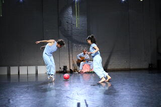 Auf dem Foto sind die zwei Performer*innen spielerisch mit einem Ball zu sehen. Im Hintergrund sitzt mittig ein Musiker mit einer Akustik-Gitarre.