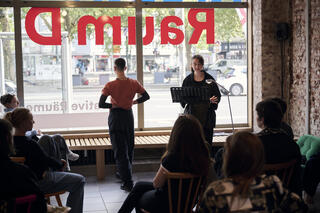 Ein Raum mit großen Schaufenster, auf das spiegelverkehrt der Schriftzug "KreativRaum D" zu lesen. Eine Gruppe von Personen sitzt auf Stühlen vor einer weiteren Person, die vor einem Notenständer steht und etwas vorliest. Links neben ihr steht ein*e Performer*in, die*der mit dem Rücken zu den sitzenden Personen zugewandt ist.