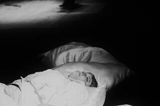 Im Vordergrund ist eine Person zu sehen die auf dem Boden liegt auf großen Kissen und schaut nach oben. Um die Person herum sind mehrere Kissen. Das Foto ist in schwarz-weiß. Im Hintergrund ist eine weitere Person zu erkennen, je doch verschwommen.