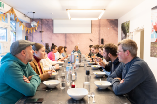 Mehrere Personen sitzen sich gegenüber an einem langen Tisch, essen und reden, Gläser und Flaschen stehen in der Tischmitte.
