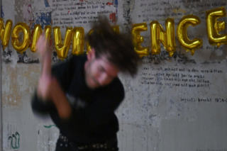 Person in Bewegung und unscharf im Vordergrund, goldene Buchstaben formen Wort an Wand mit Textfragmenten im Hintergrund.